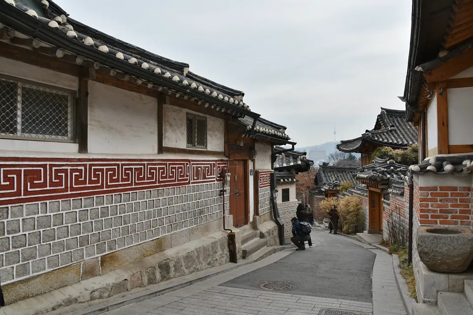 Narrow Bukchon lane sloping down between hanok houses with red patterned walls, a hazy city skyline beyond.